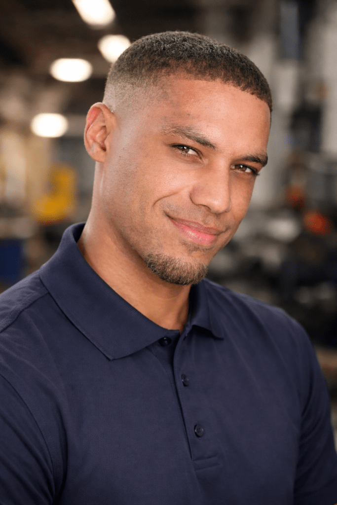 A close-up portrait of a smiling man with short hair, wearing a navy blue polo shirt, set against a blurred industrial background.